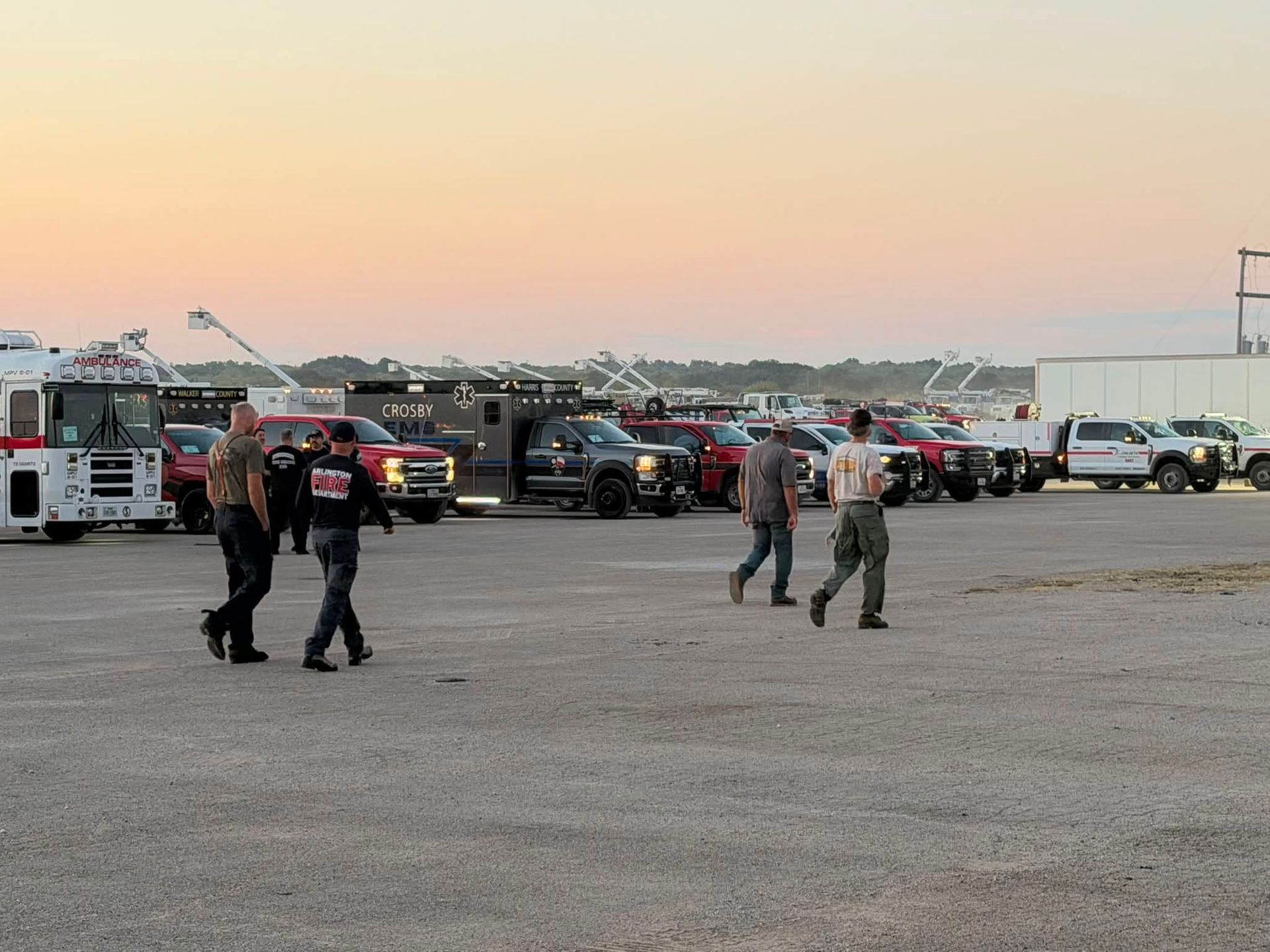 People walking across a gravel lot with parked emergency vehicles at sunset.