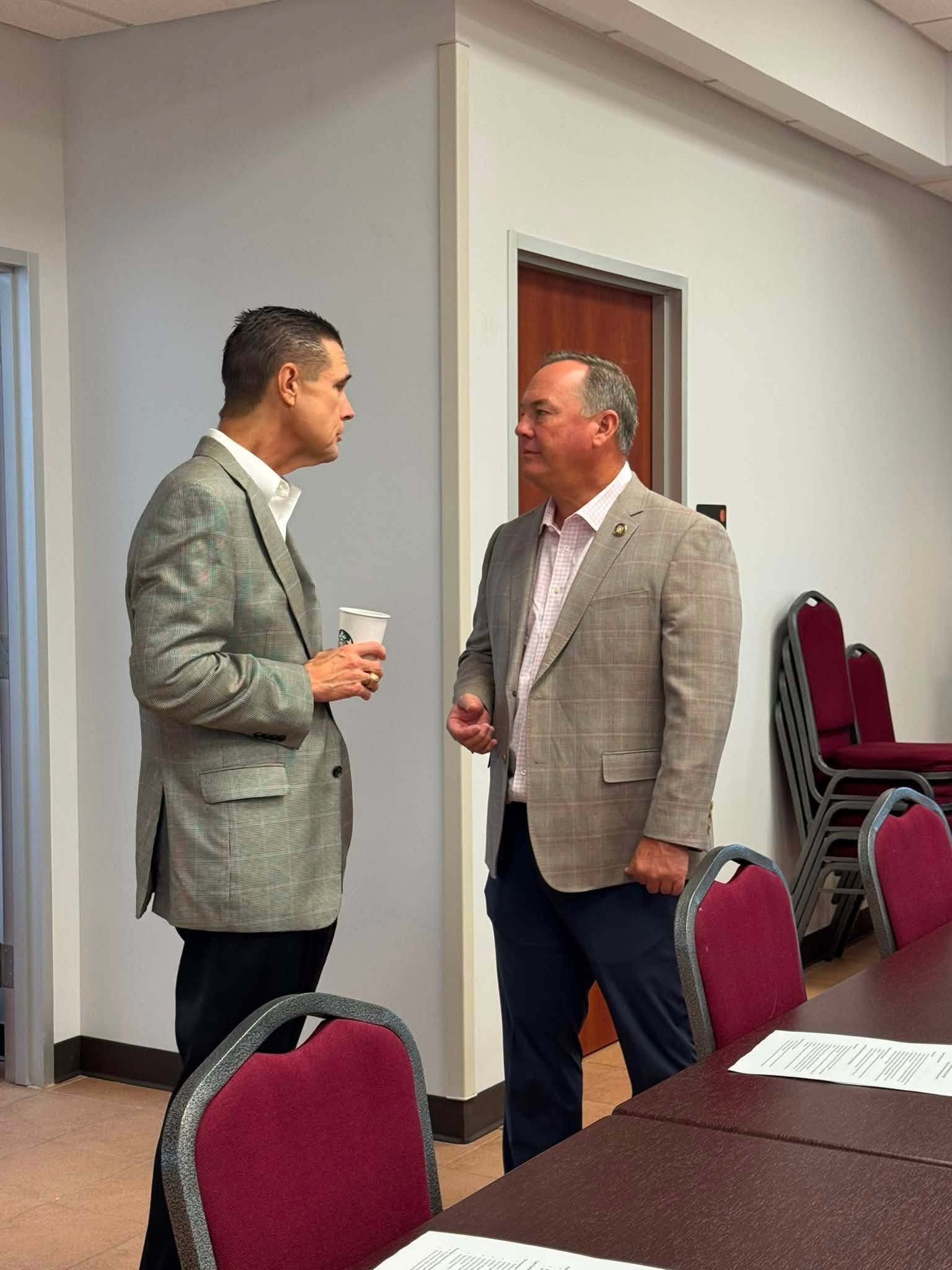 Two men in blazers chat beside stacked chairs in a conference room.