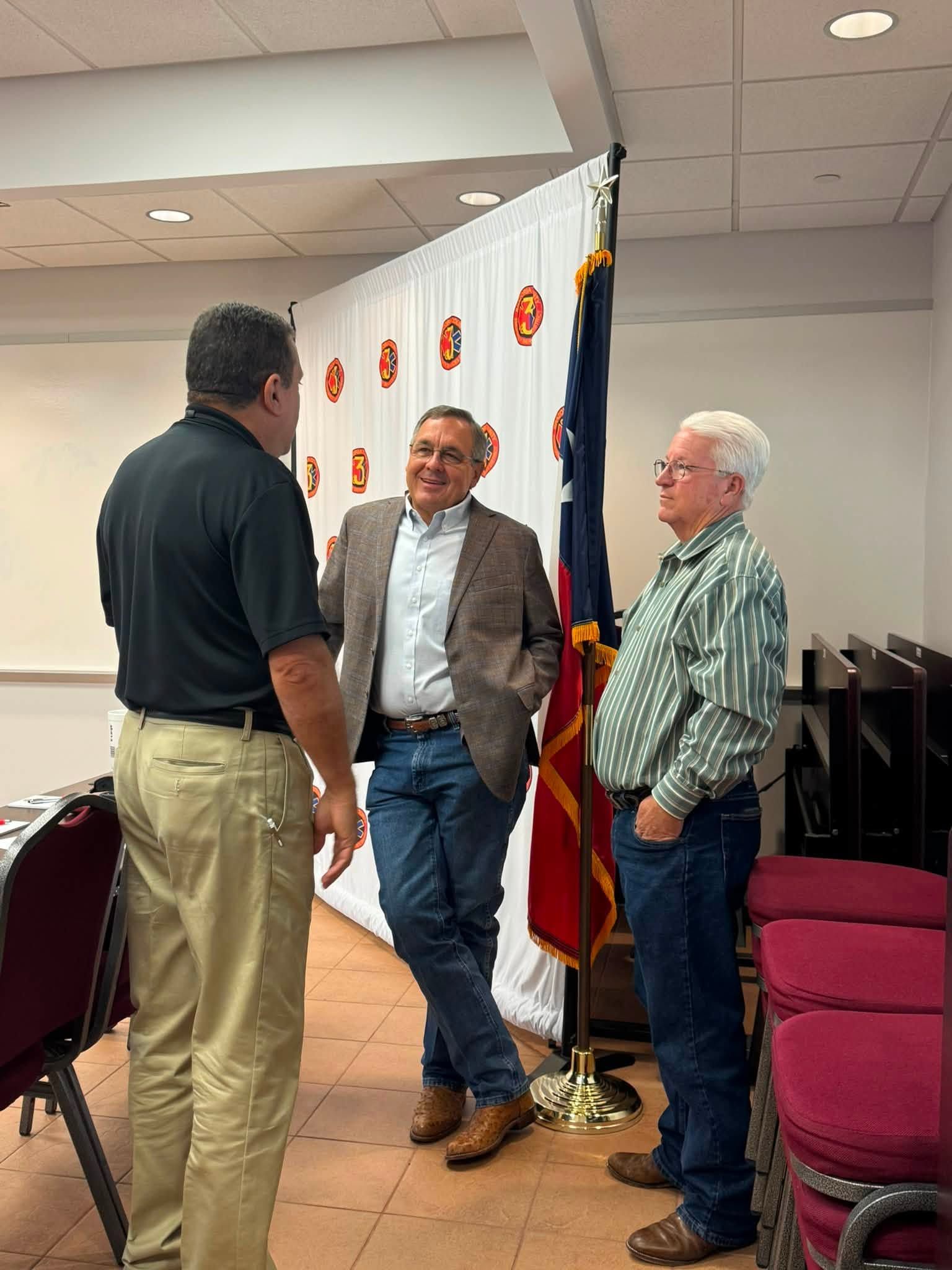 Three men chatting beside a U.S. flag in a meeting room with chairs and a banner backdrop