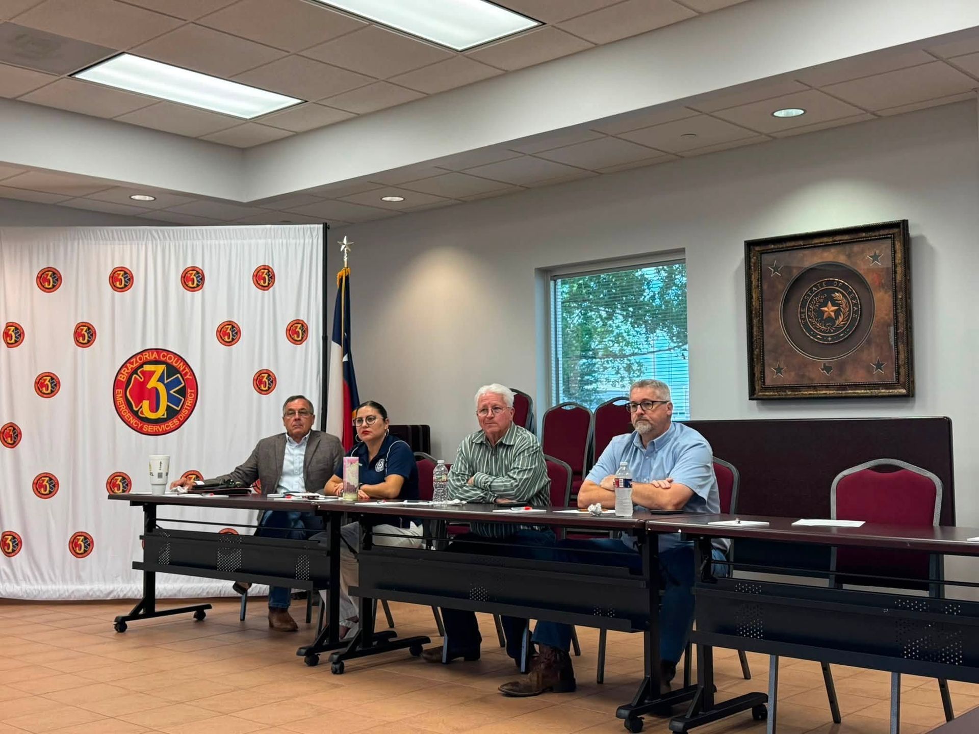 Panel discussion in a meeting room with four seated speakers at a long table and a backdrop banner and flag