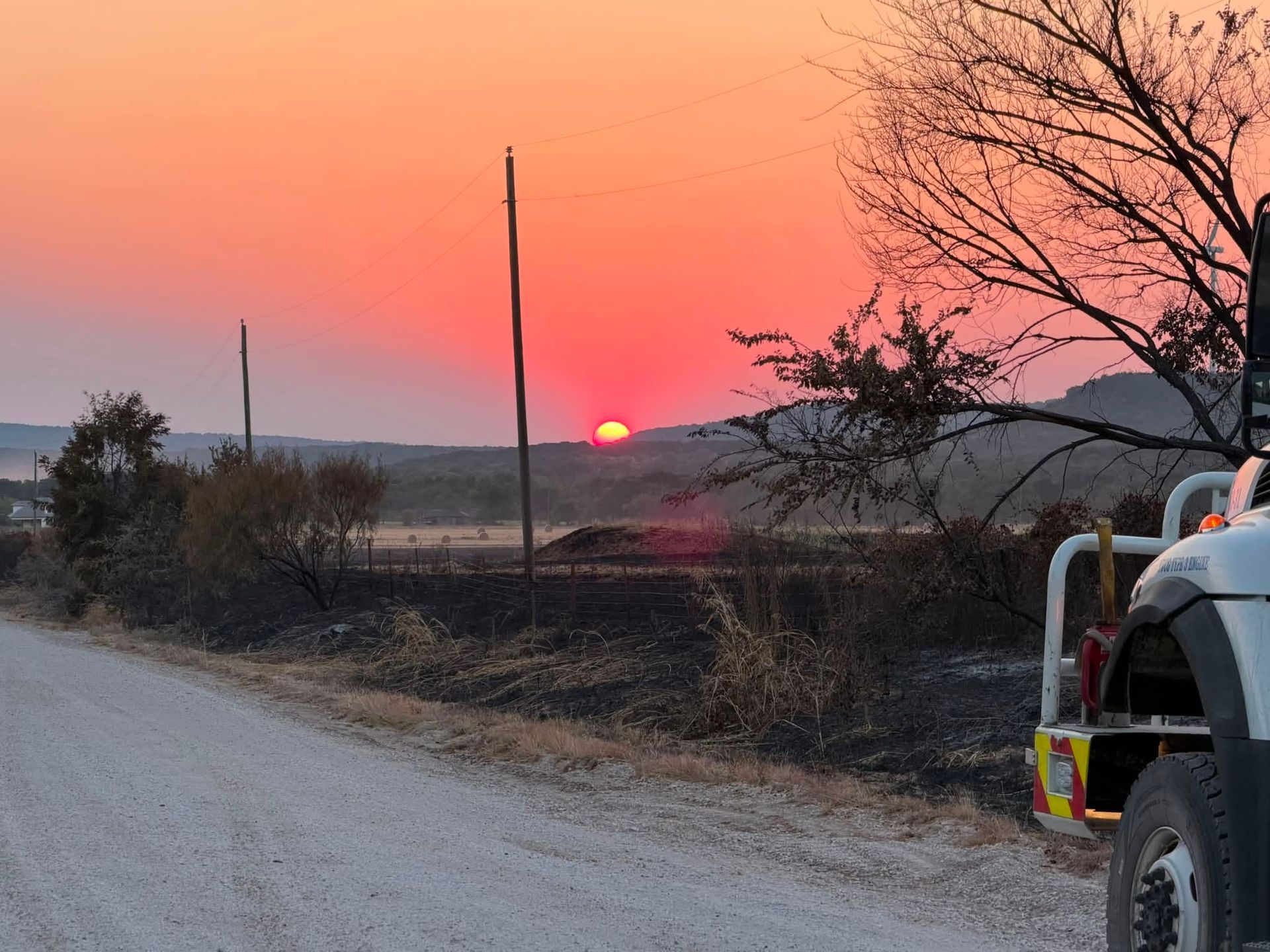 Sunset over a rural dirt road, with a truck at right and bare trees silhouetted against a pink-orange sky