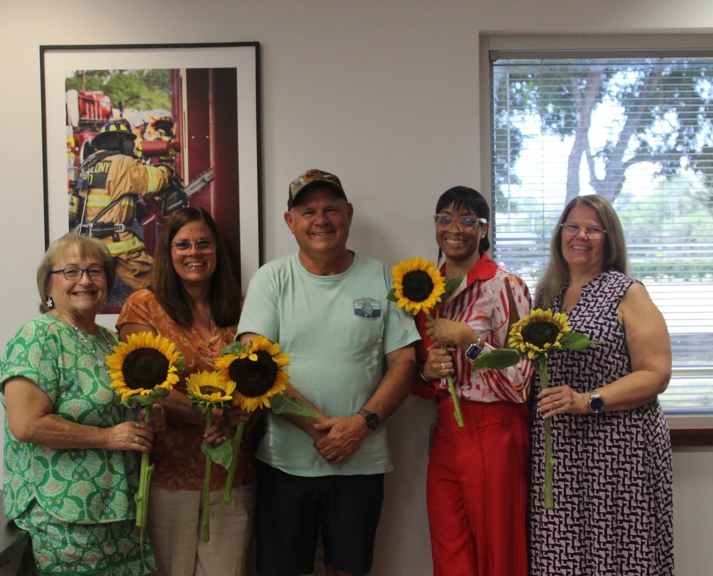 Five people holding sunflowers stand smiling in an office or lobby with framed art and a window behind them.