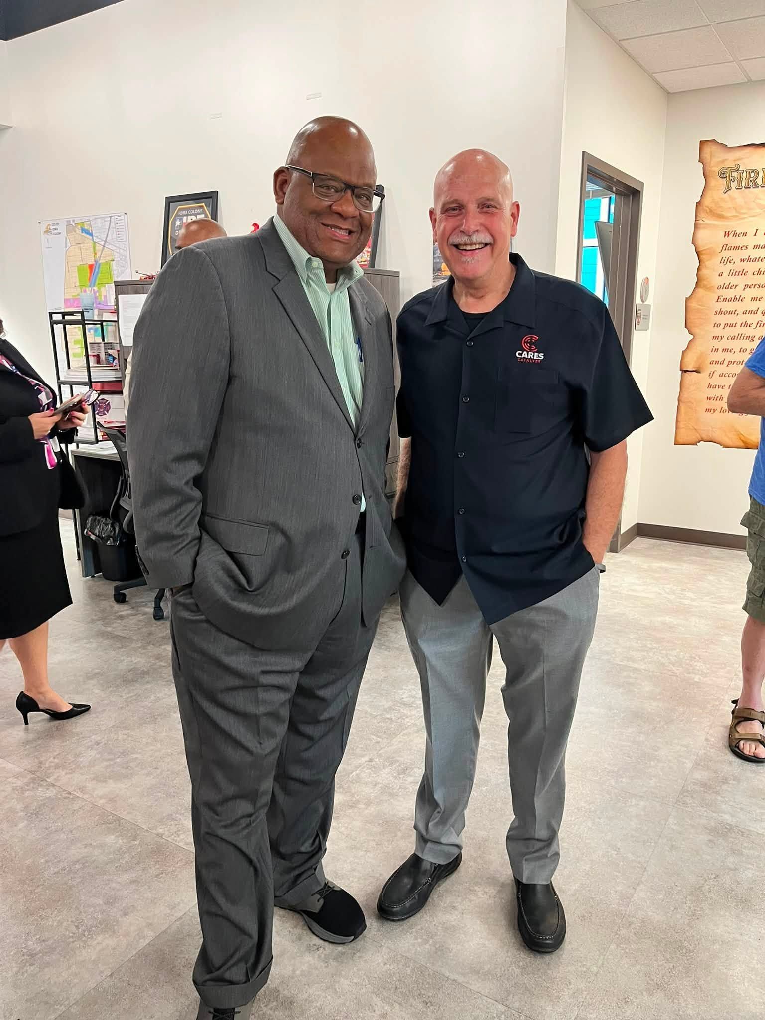 Two men smiling and posing together at an indoor event, one in a gray suit and one in a navy polo shirt.