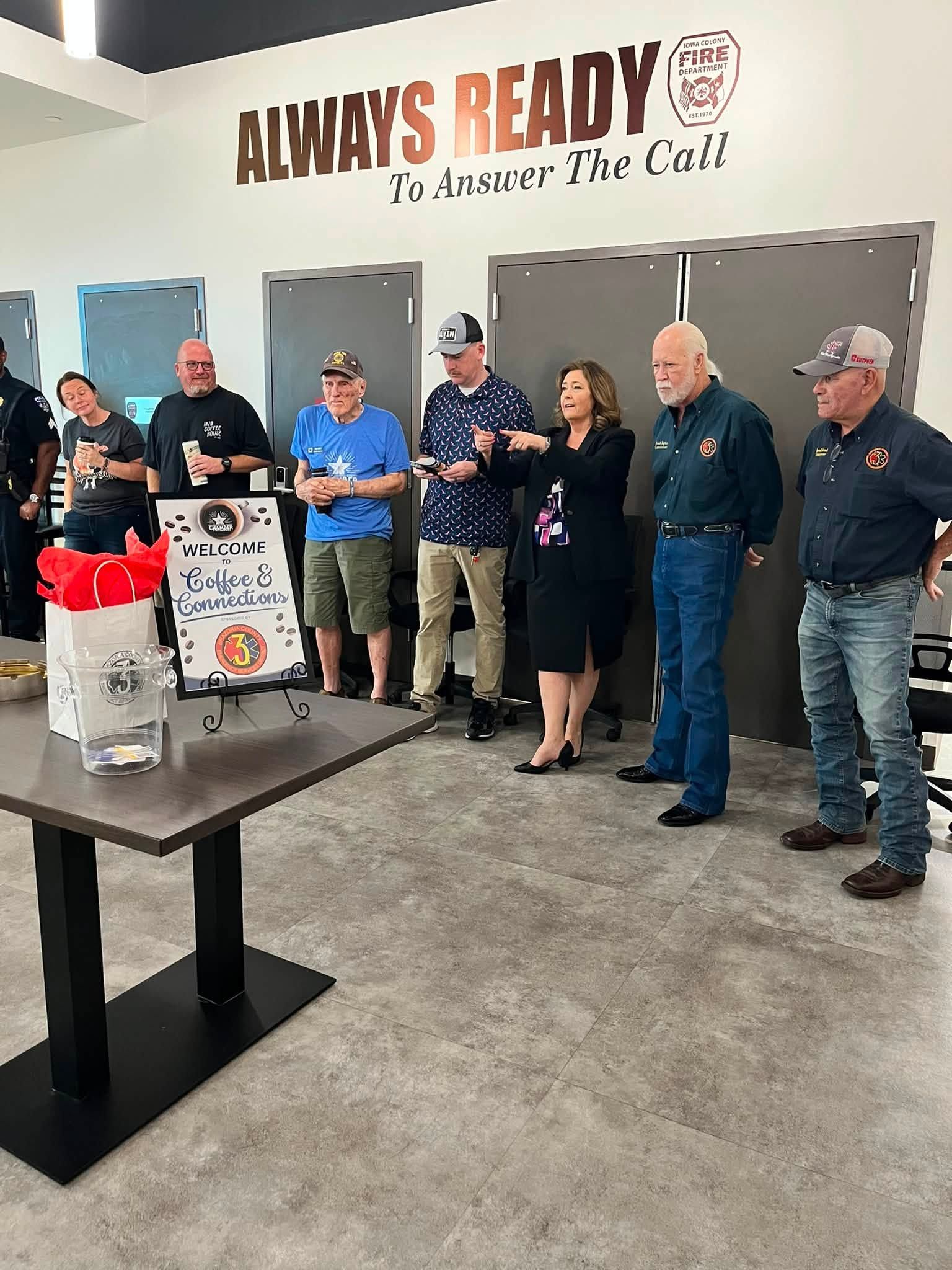 People standing together at a community gathering inside a room with a wall sign reading “Always Ready.”