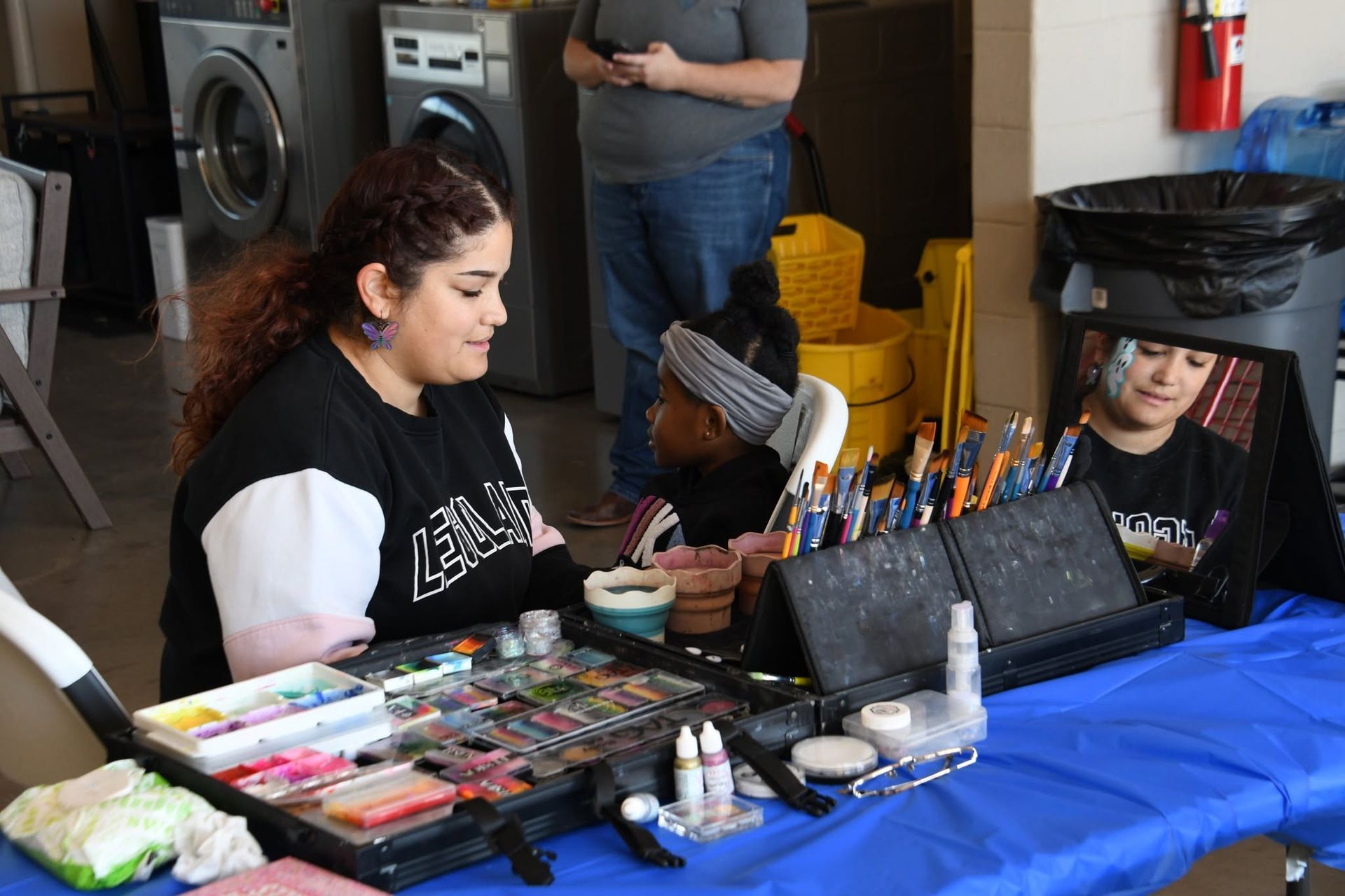 Artists seated at a table with makeup palettes and brushes under a blue tablecloth, preparing face painting supplies