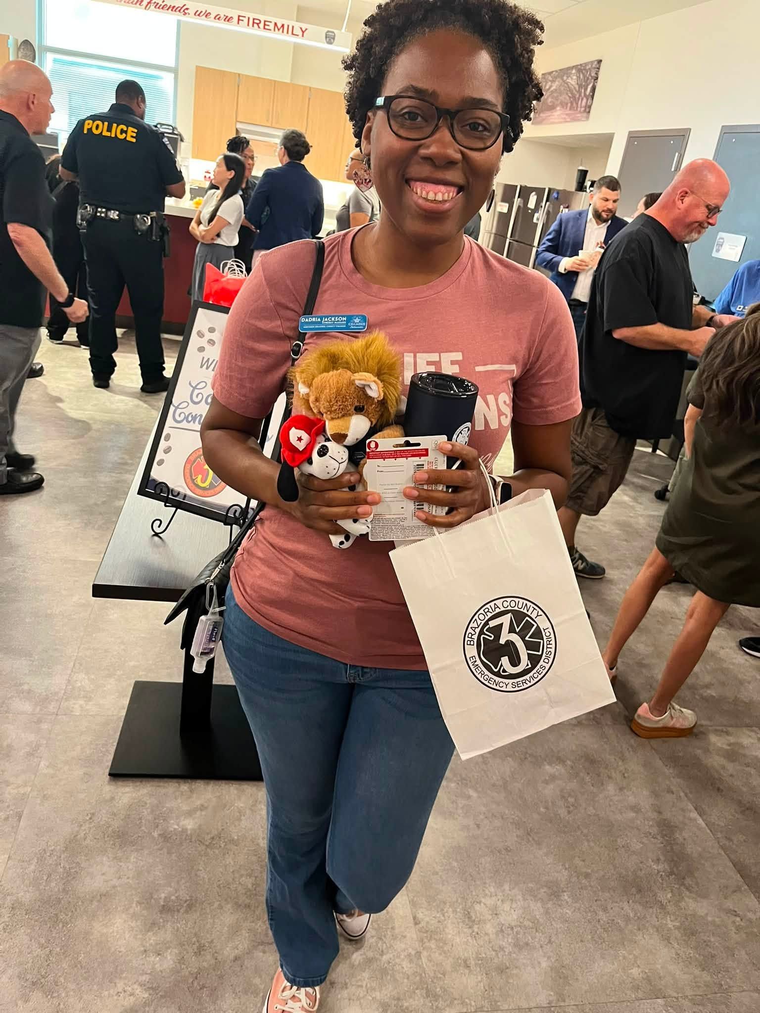 Woman smiling, holding a stuffed toy and drinks at an indoor event, with a tote bag over her arm.