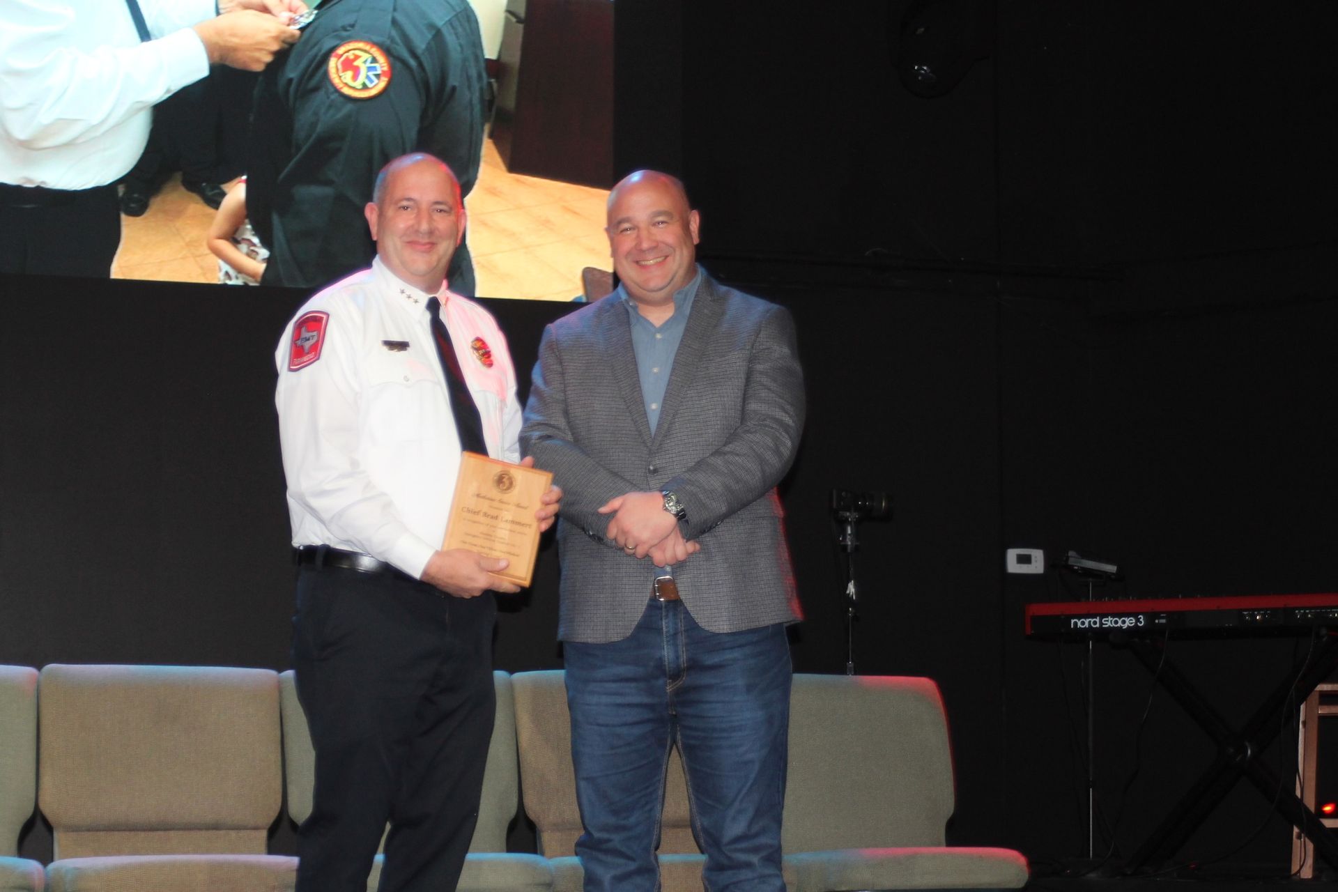 Two men on stage; one in an EMS uniform holds a plaque, the other in a blazer smiles. They stand in front of a dark backdrop.