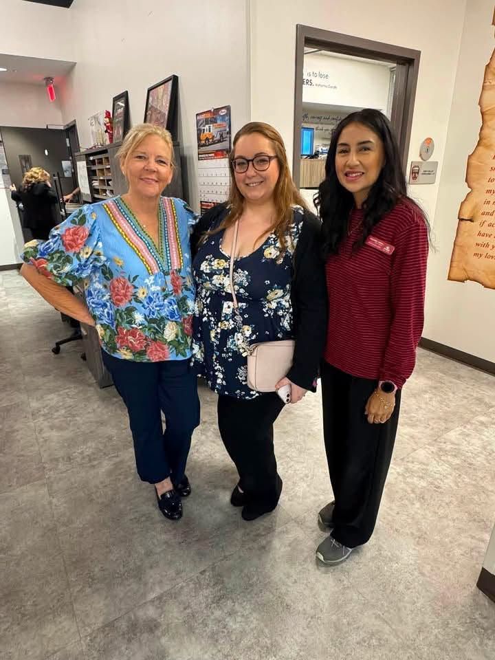 Three women smiling indoors, wearing colorful blouses and standing in a hallway.