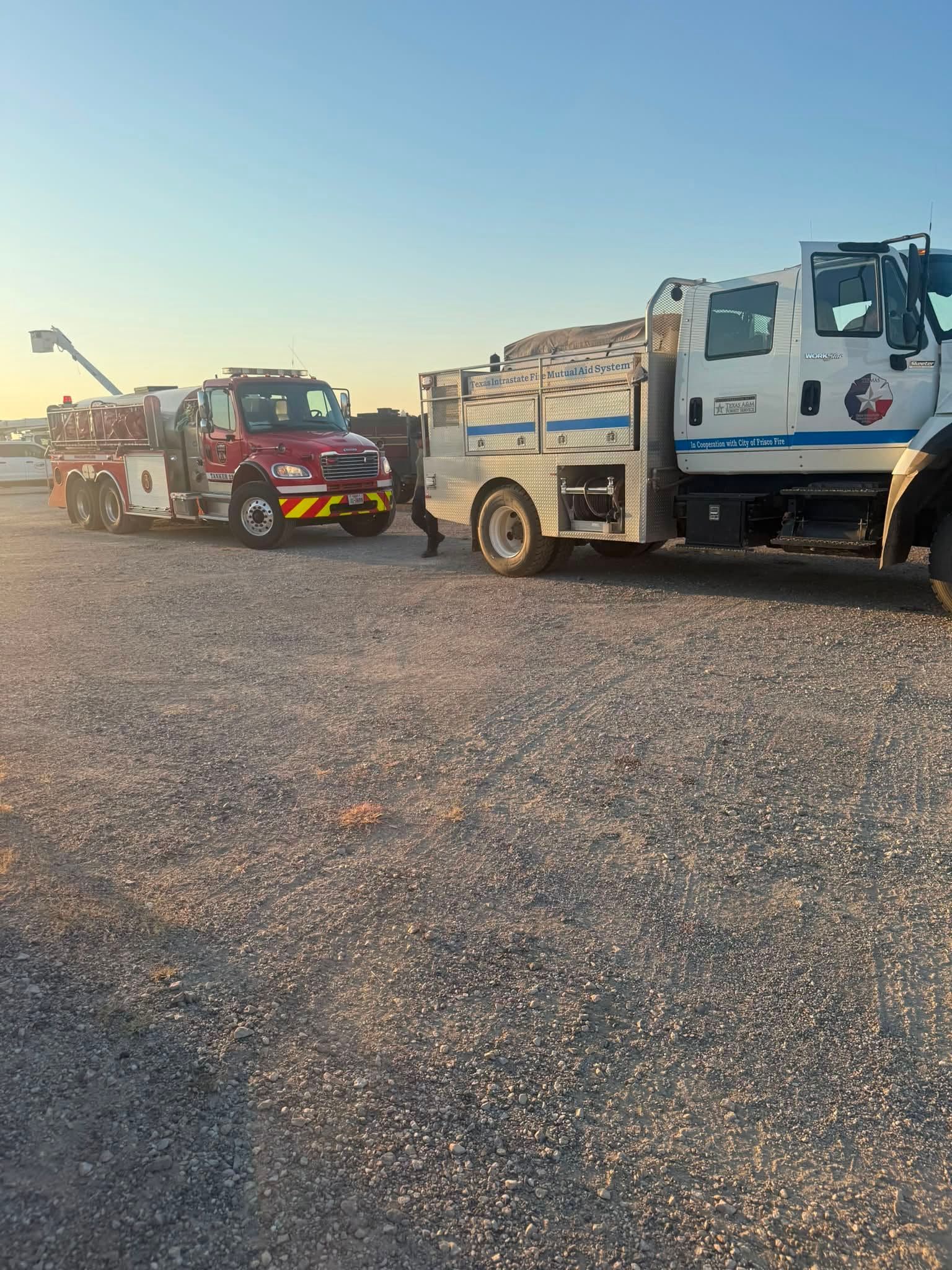 Two fire apparatus parked on a gravel lot at sunset, one red and one white-blue.