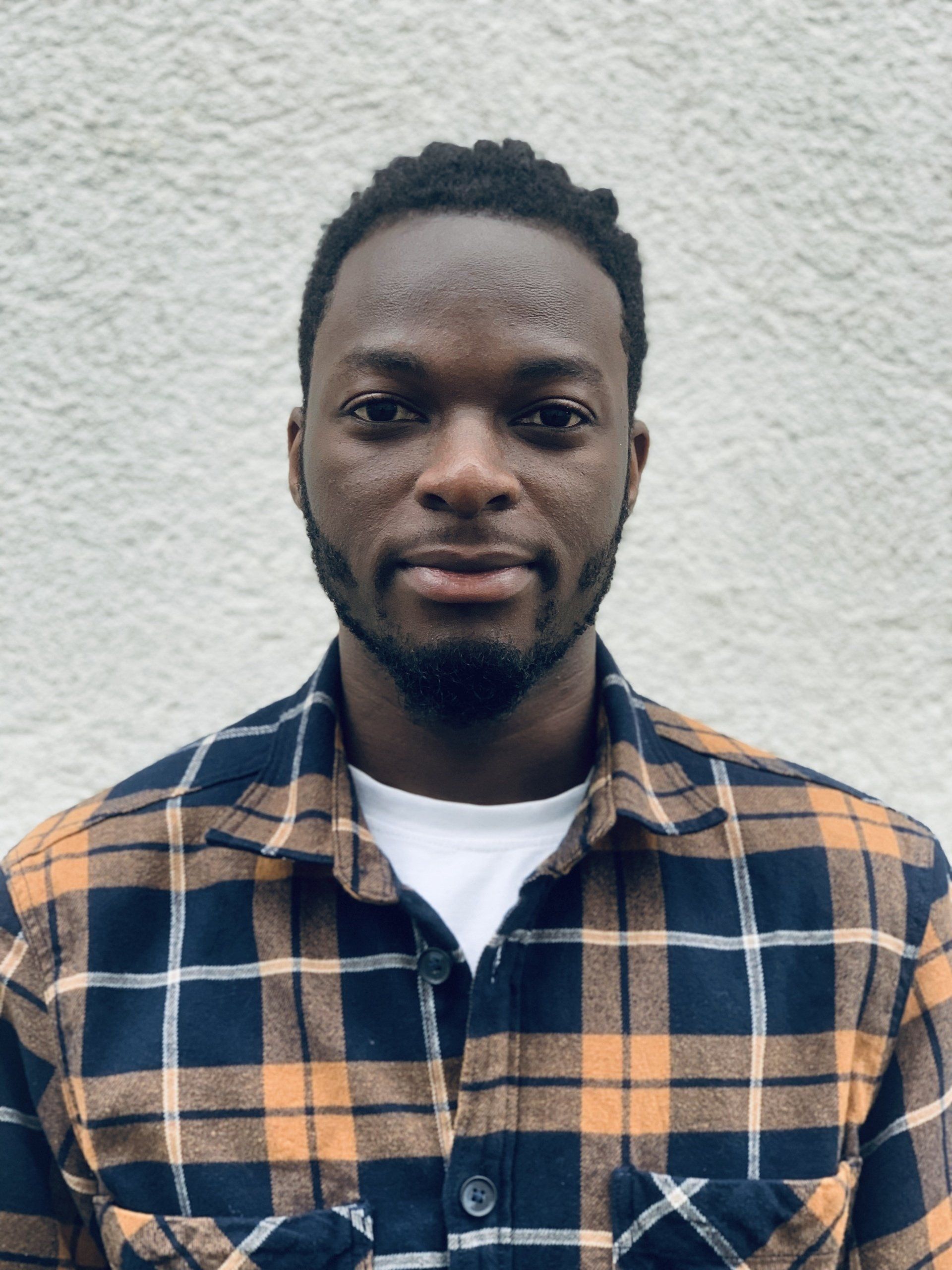 A man wearing a plaid shirt is standing in front of a white wall.