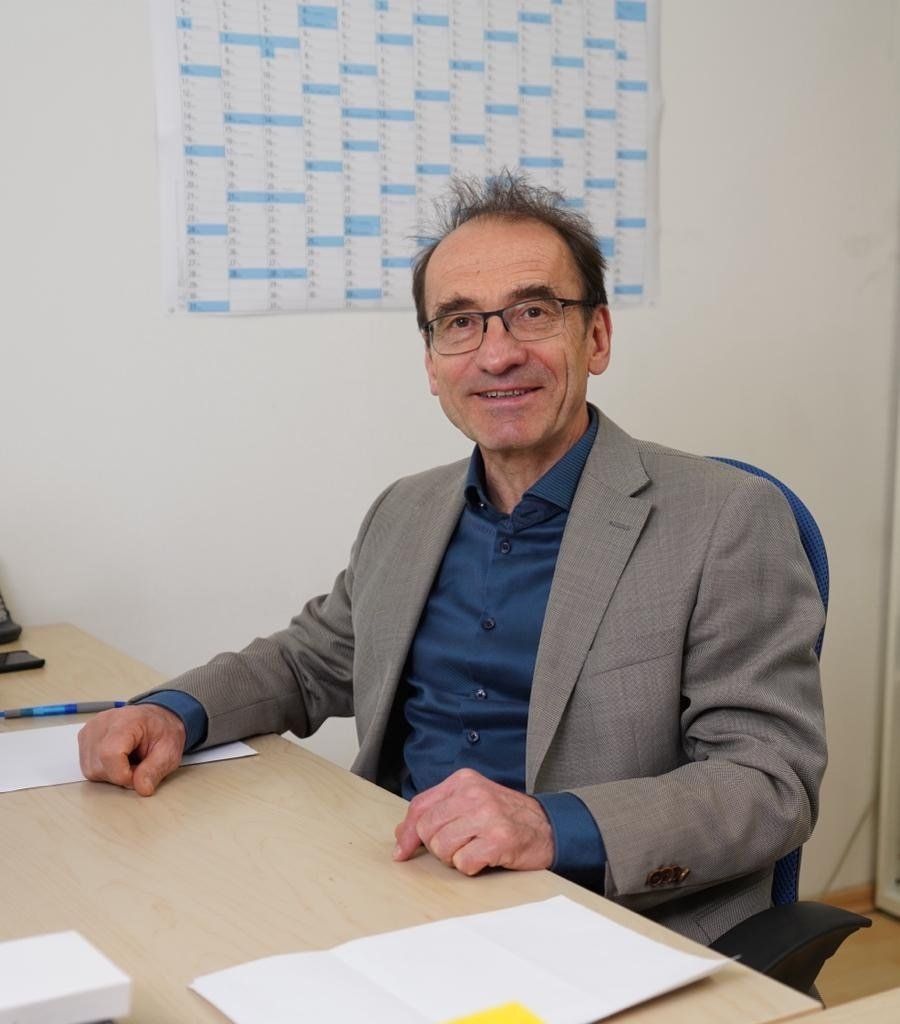 A man in a suit is sitting at a desk with a calendar on the wall behind him
