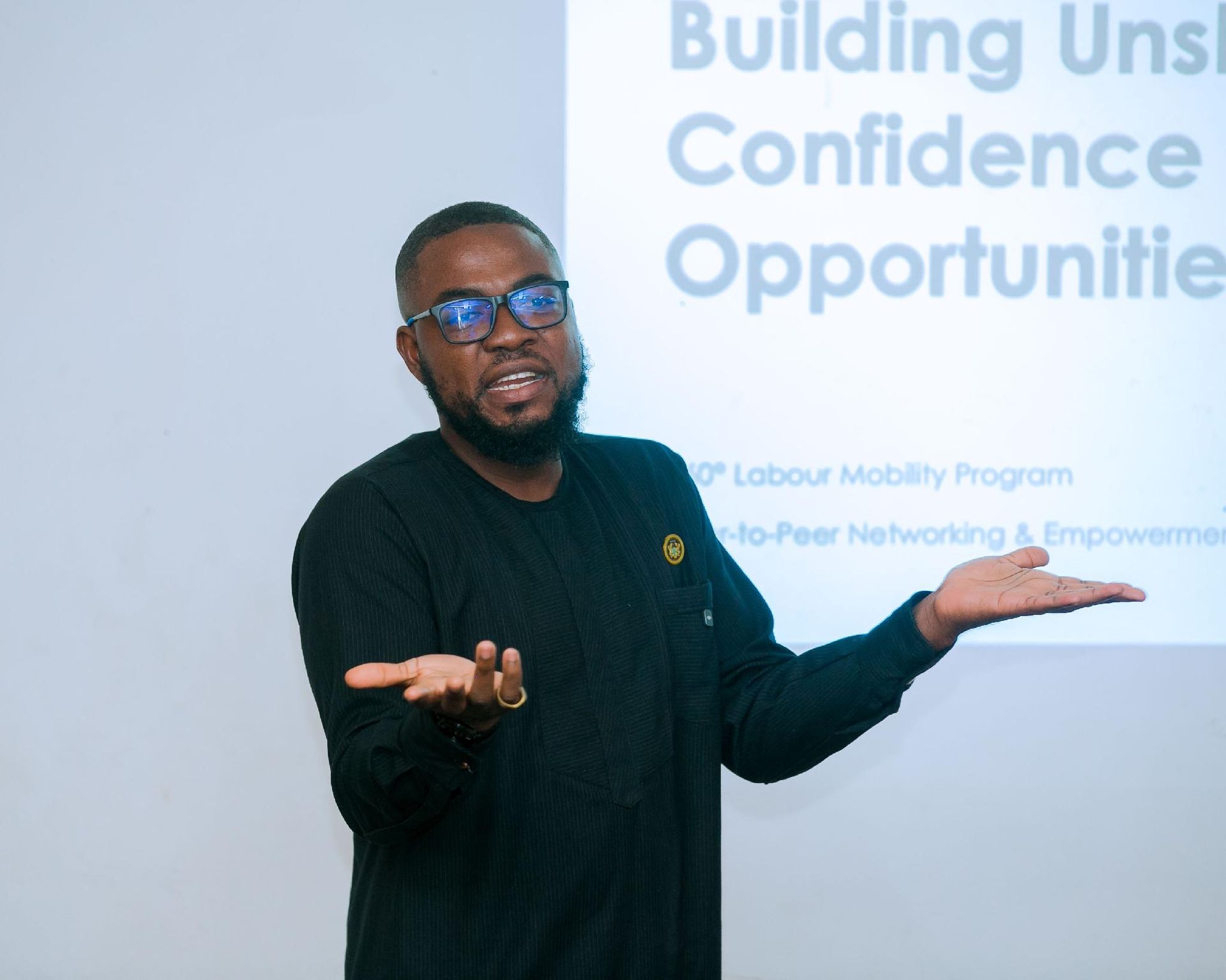 Man in dark shirt and glasses presenting before a slide, hands raised