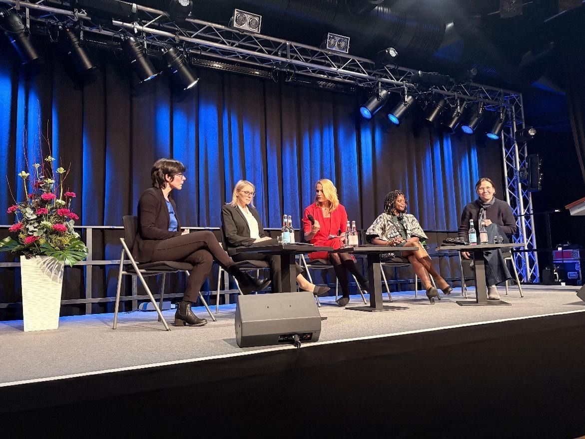 Panel discussion on a stage with five people seated at a table, blue backdrop.