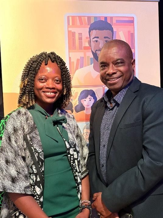 Two people smiling in front of a poster. Woman in green dress and patterned jacket; man in a blazer.