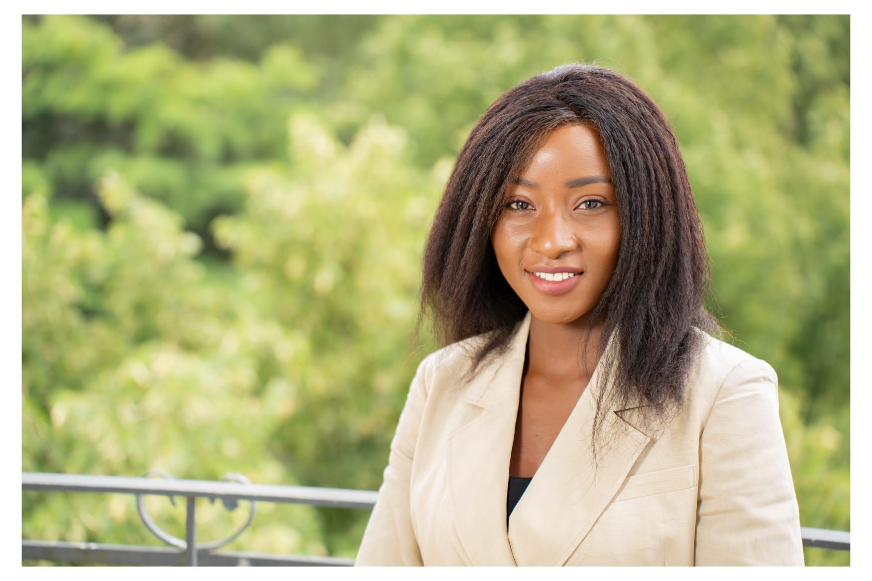 A woman in a tan jacket is standing on a balcony with trees in the background.