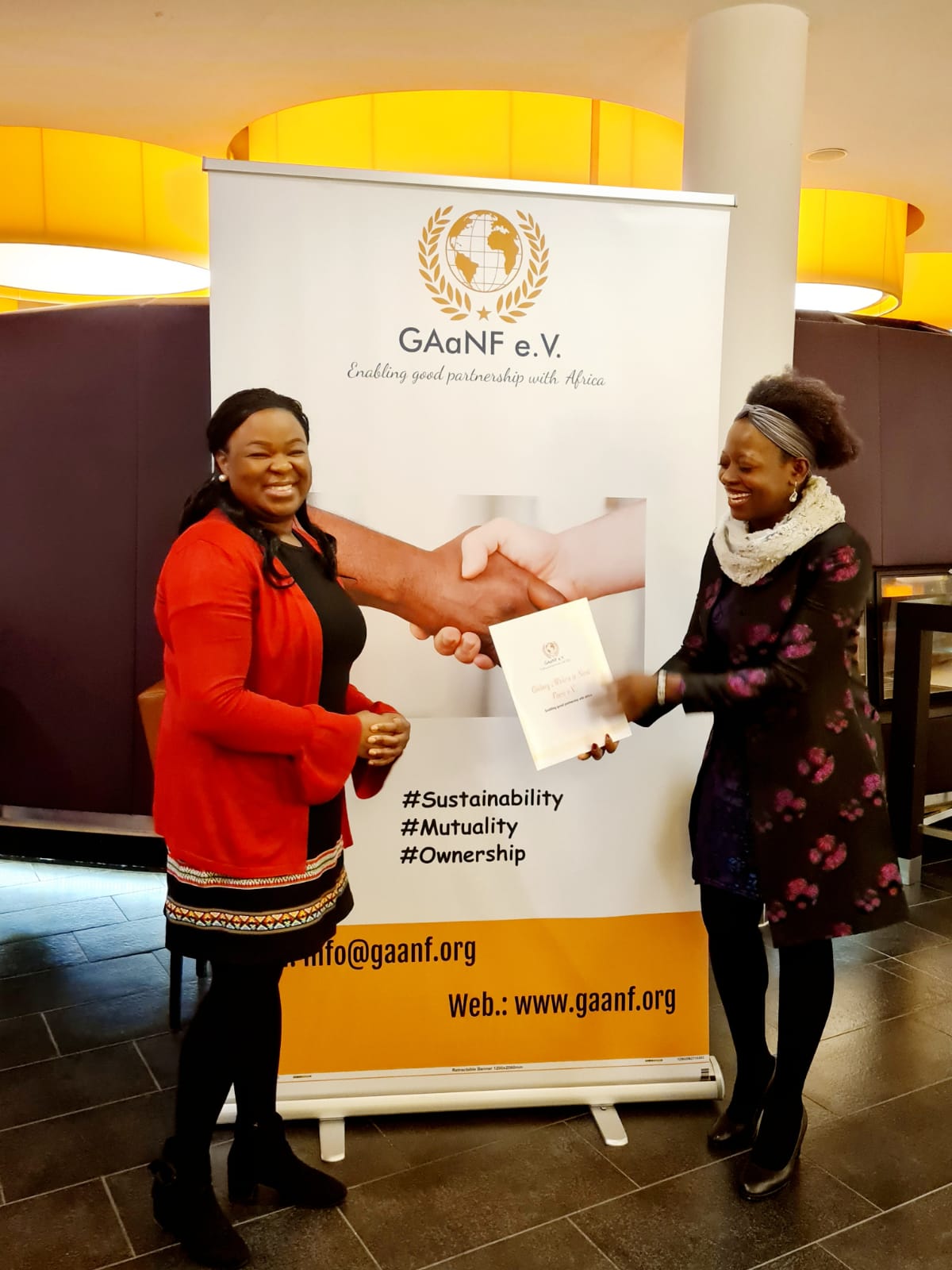 Two women are shaking hands in front of a sign.