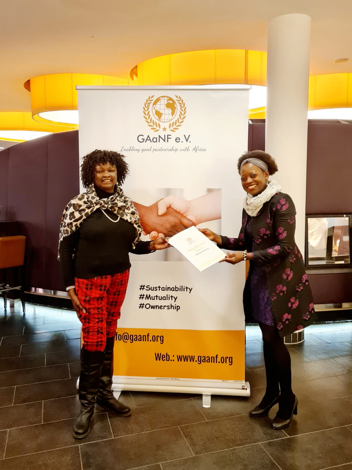 Two women are shaking hands in front of a banner.