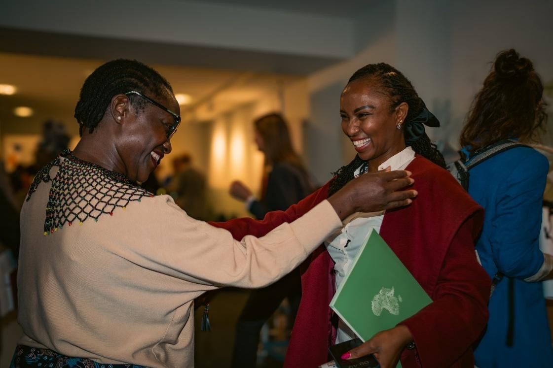 Two smiling women embrace indoors; one holds a green folder. Warm lighting, blurred background.