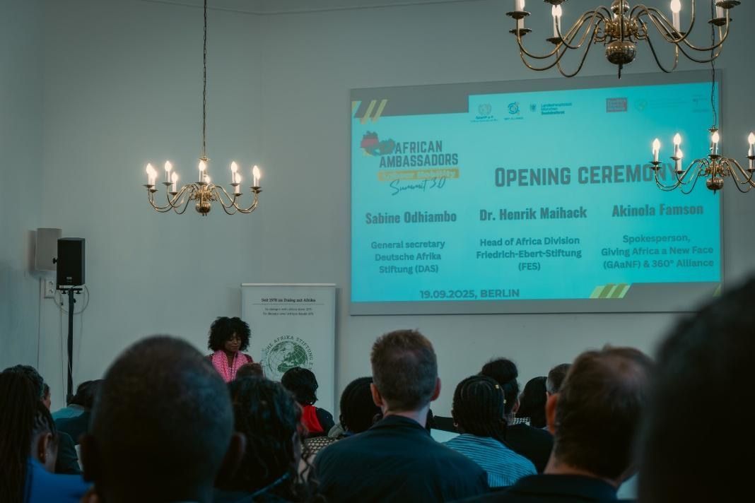 People at a conference listening to a speaker in a room with chandeliers and a screen.