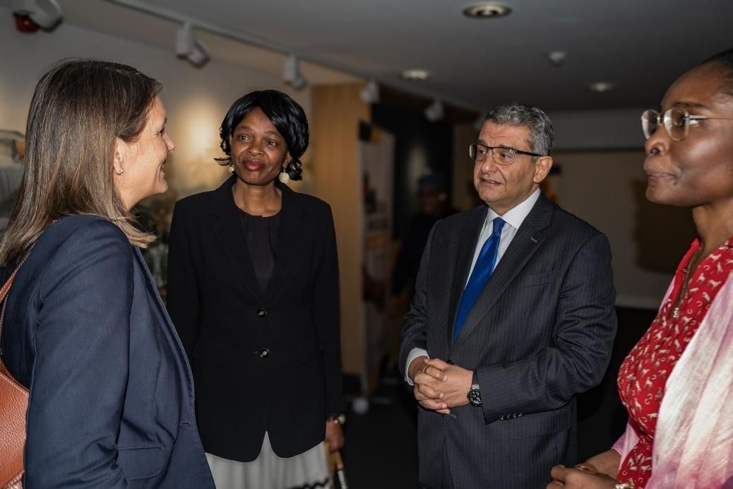 Four people in suits conversing in an indoor setting. One woman in red, others in black or blue.