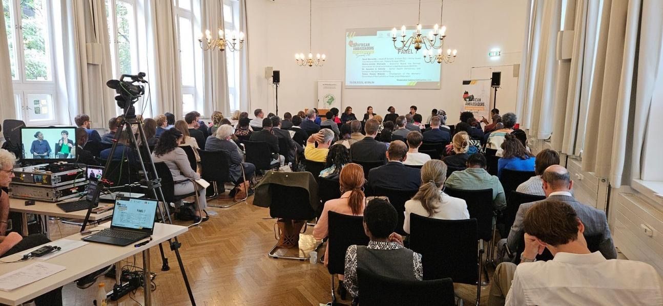 Conference in a large room, attendees seated, screen at front, video recording in progress.