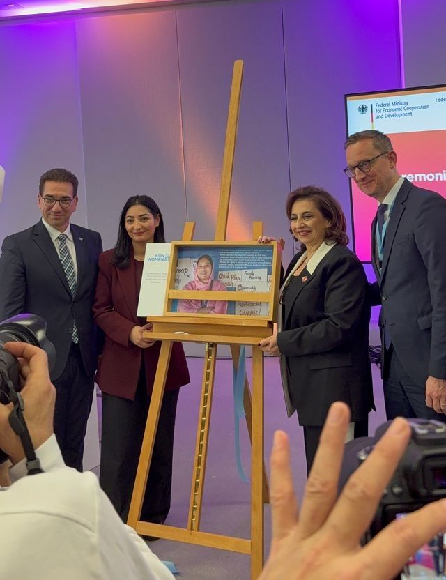 Four people holding a framed photo on an easel, indoors, at a ceremony. People smiling, and a person taking a photo.