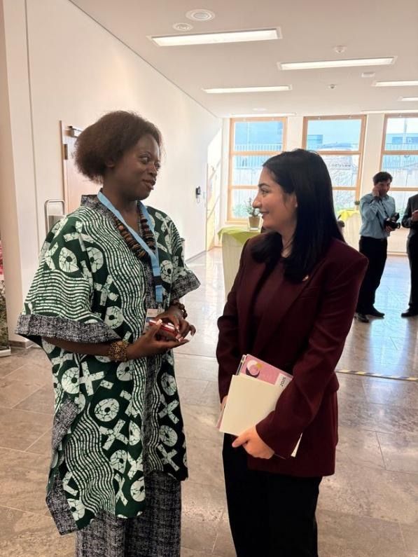Two women in conversation in a hallway. One wears a patterned dress, the other a blazer, smiling. Bright setting.