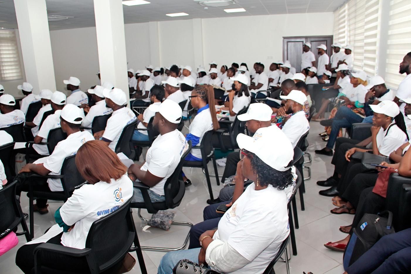 Large group seated in a meeting room, many wearing white caps and shirts, facing forward.
