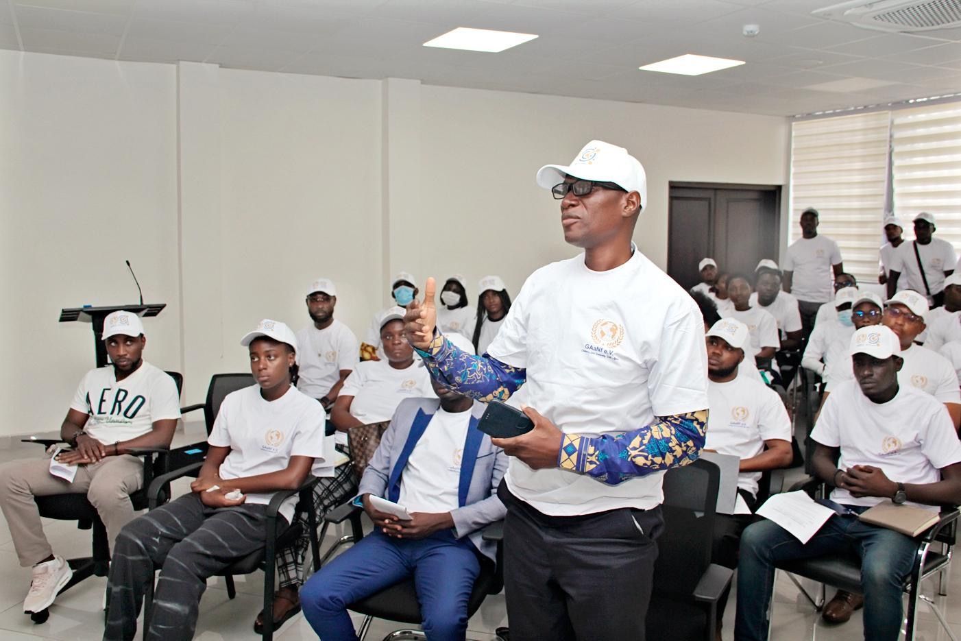 Man in white cap and shirt stands in a meeting room, holding blue items, with seated attendees behind him.