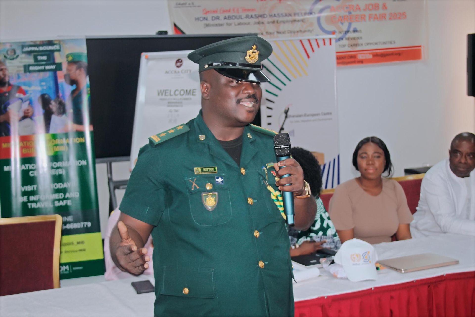 Uniformed officer speaking into a microphone at a table event, with two seated attendees behind him.