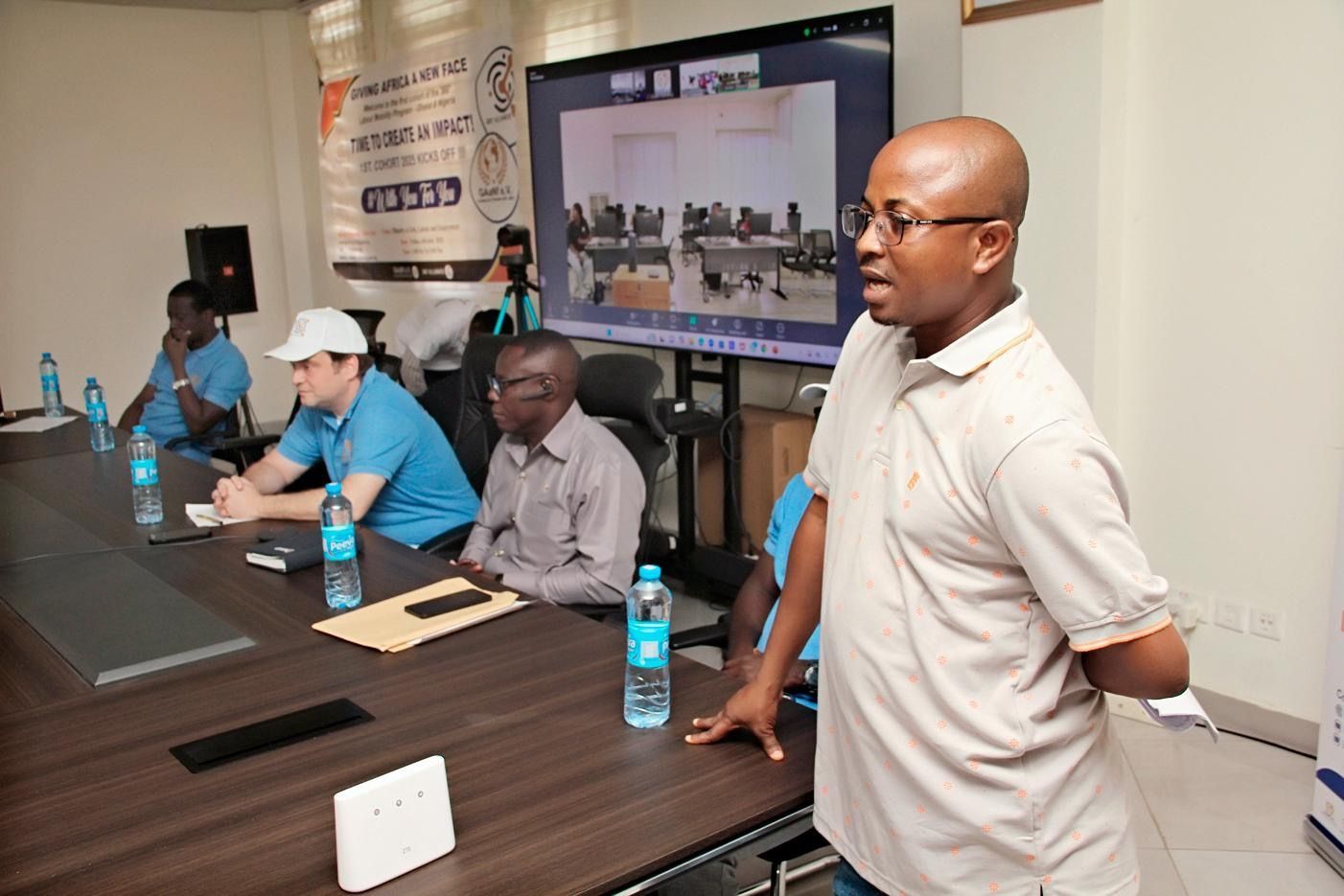 Meeting room with four people seated at a conference table and one person standing and speaking by a wall screen.