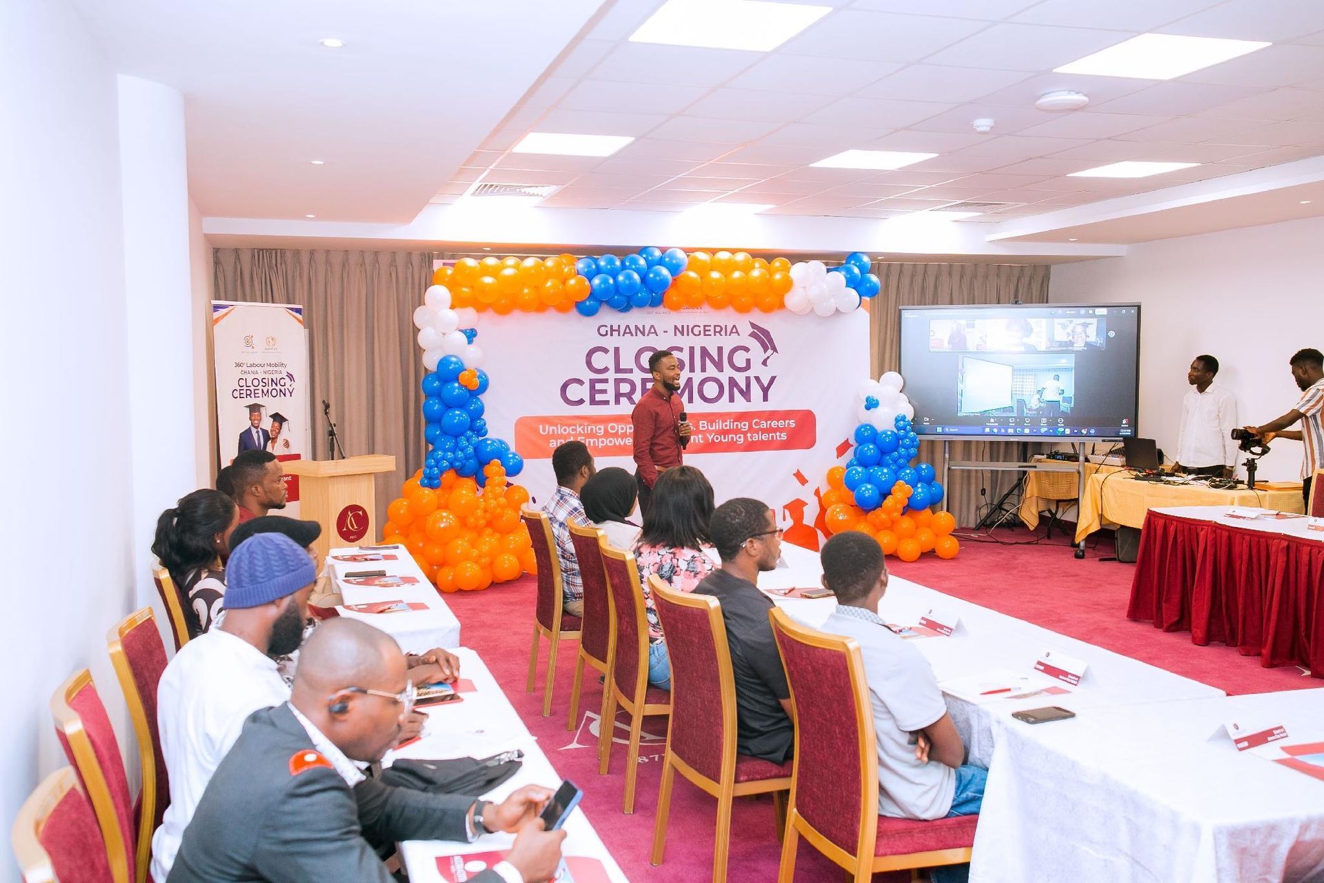 Conference hall with seated attendees facing a “Closing Ceremony” stage decorated with orange and blue balloons.