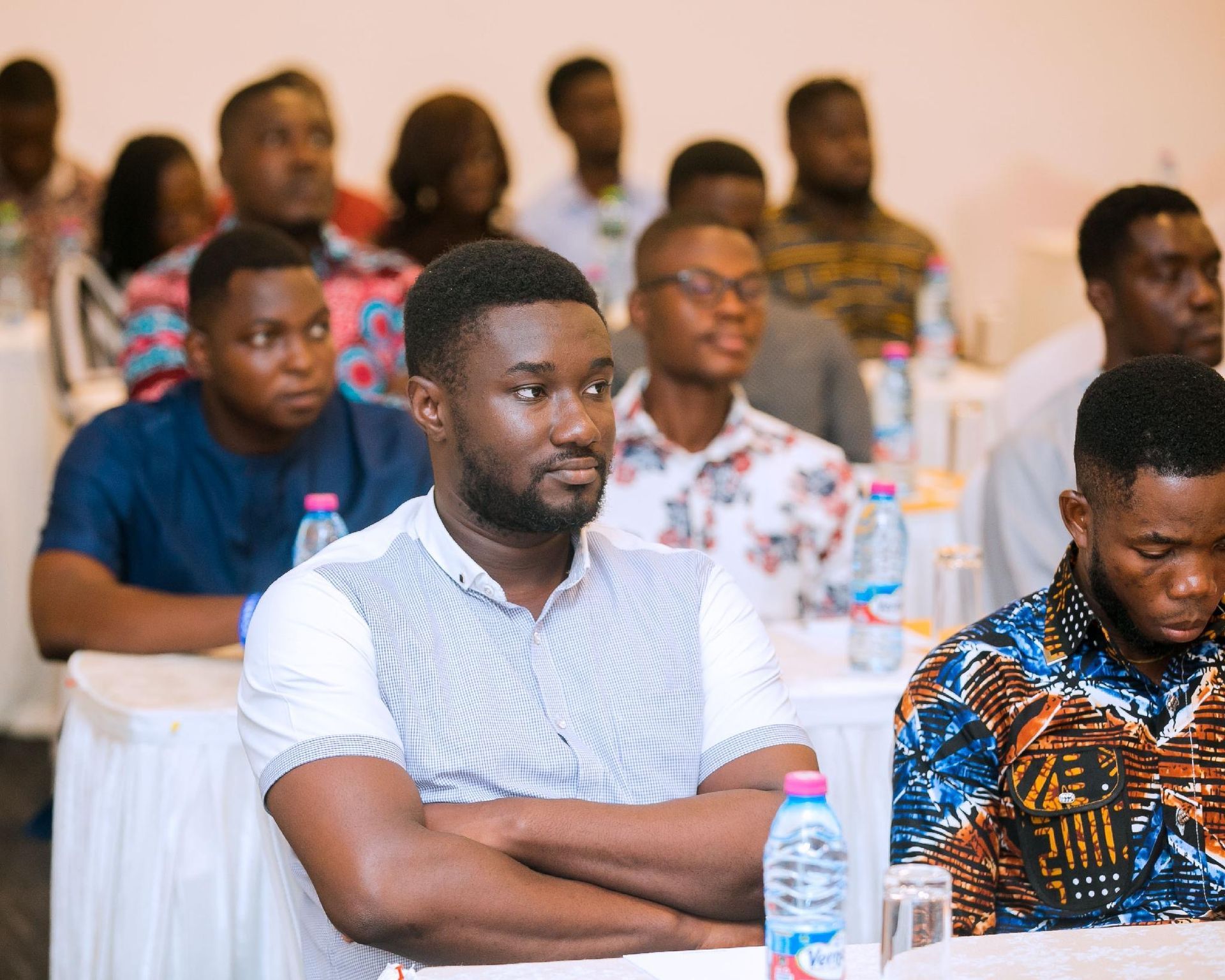 Audience seated at a conference, listening attentively at tables with water bottles and notebooks.