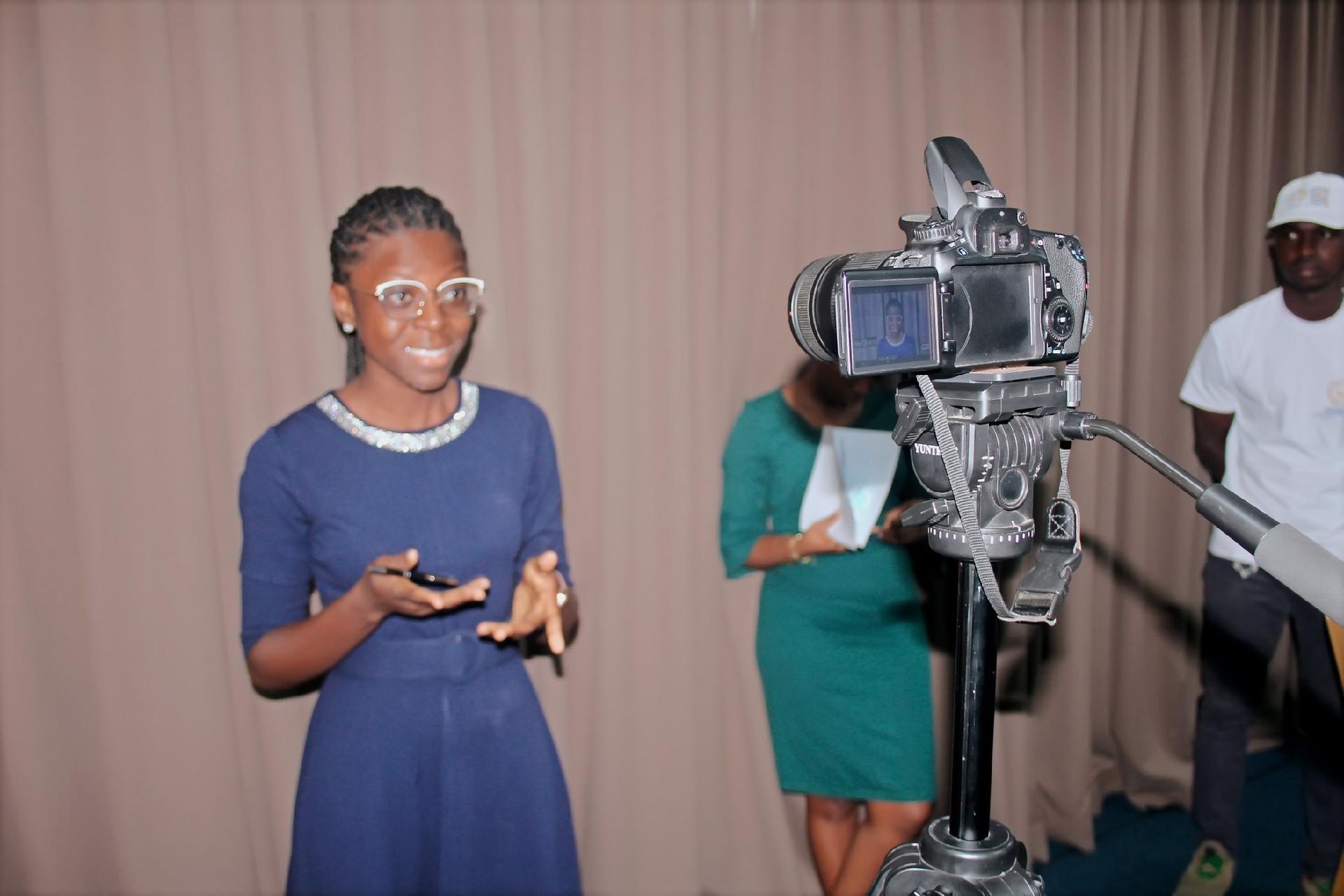 Person speaking beside a video camera in a studio, with two others standing behind a curtain.