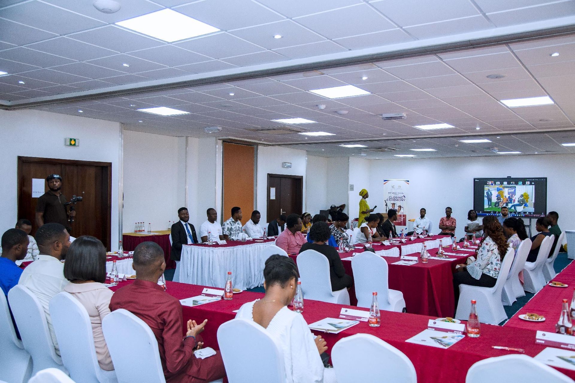 Conference room meeting with people seated around red-covered tables and a presentation screen at the front