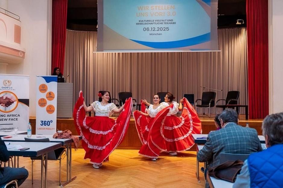 Dancers in red skirts perform on a stage during an event. People watch from tables.