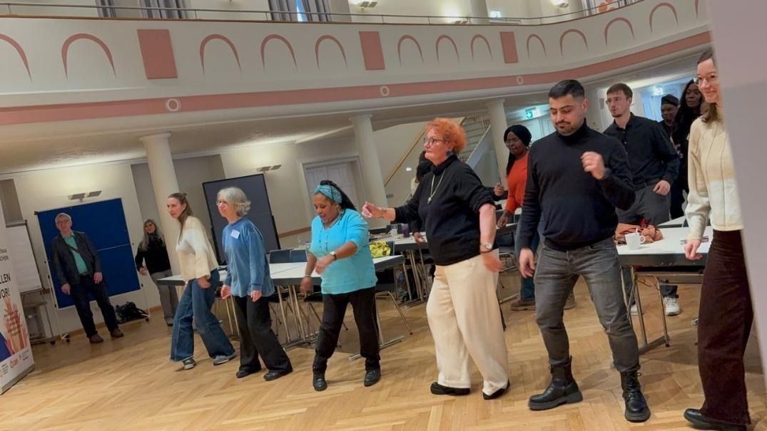 People dancing in a brightly lit room with tables and a blue backdrop.