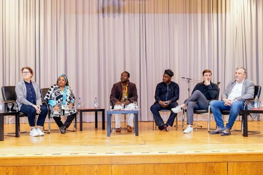 Panel seated on stage in front of a curtain. People sit at a table; they are speaking.