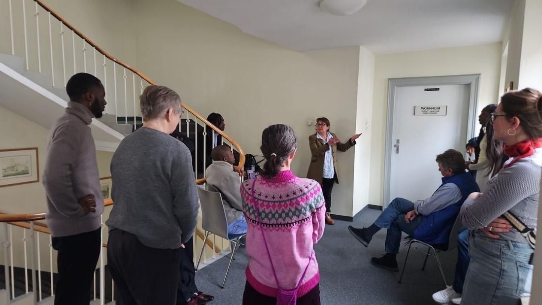 People listen to a presentation in a hallway. The speaker gestures, others look on, some sit. Staircase to the left.
