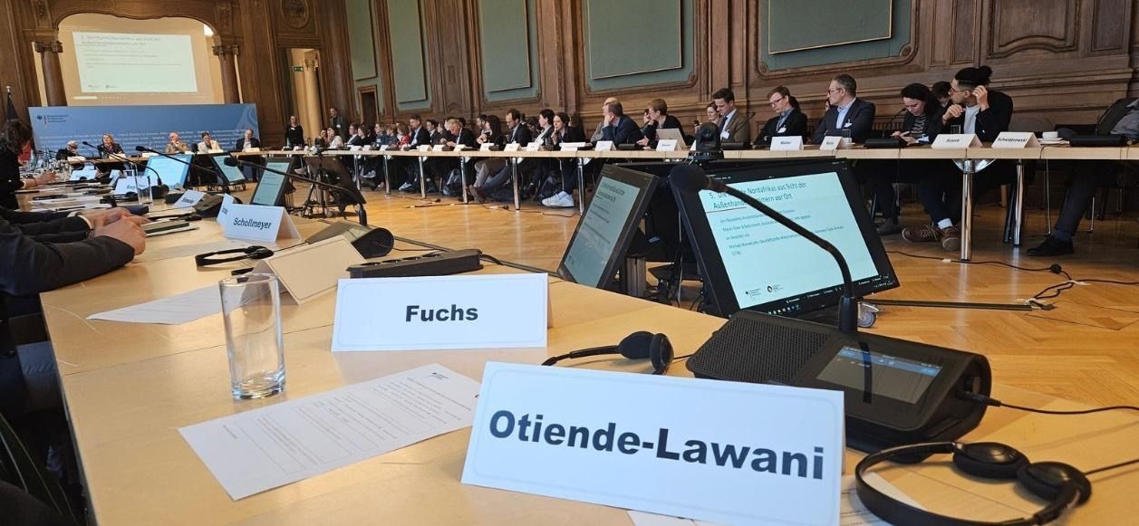 Conference room with participants seated at a long table. Names on signs. Projector in the background.