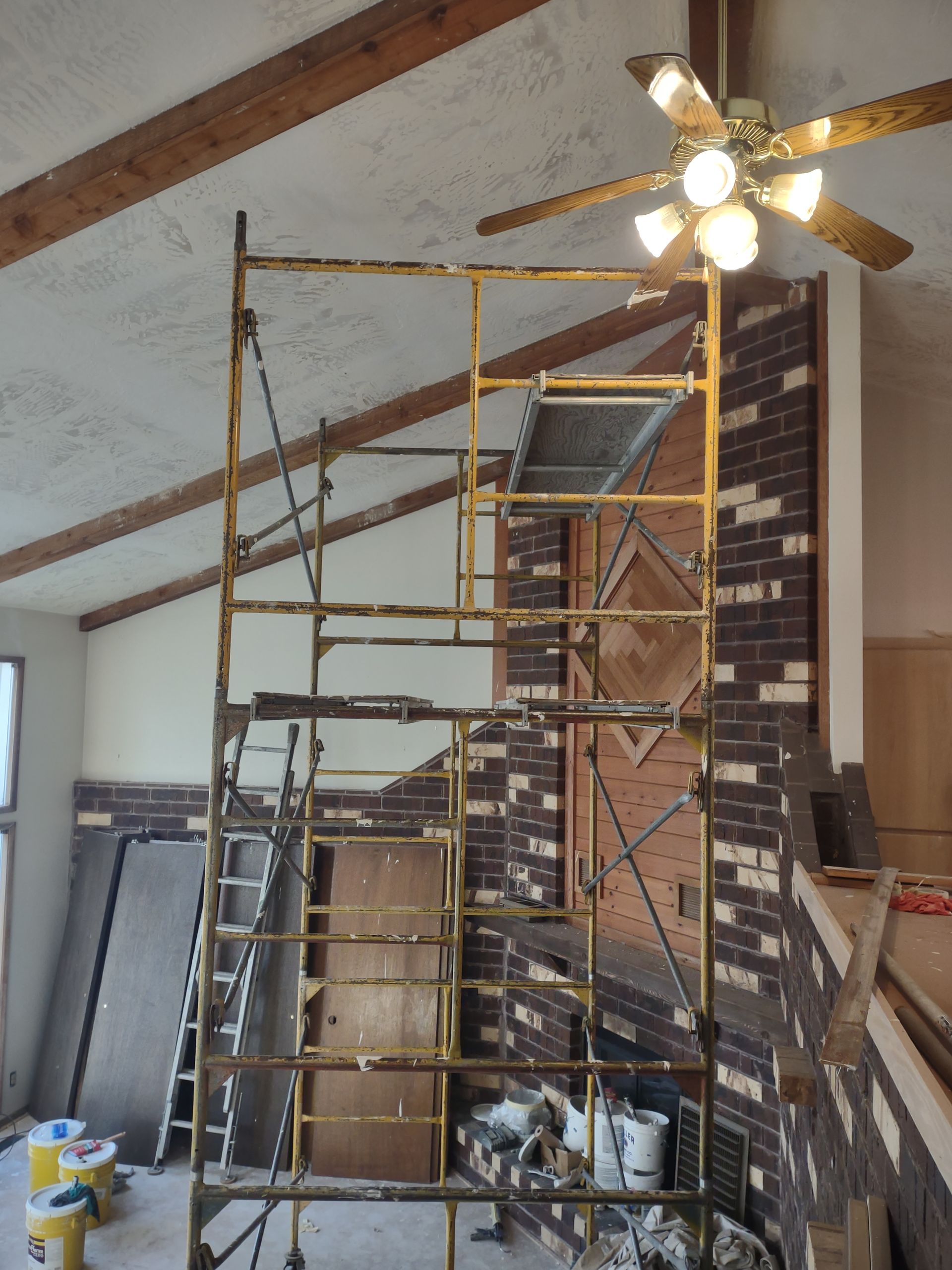 Scaffolding inside a room with exposed brick fireplace and vaulted ceiling under a ceiling fan.