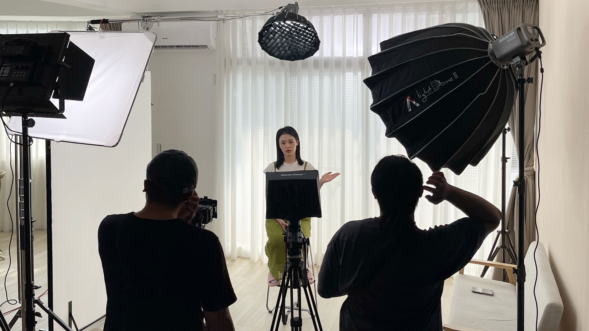 兩人正在攝影棚拍攝一位女士,棚內有燈光。女士站著,做著手勢,穿著黑色上衣和綠色褲子。