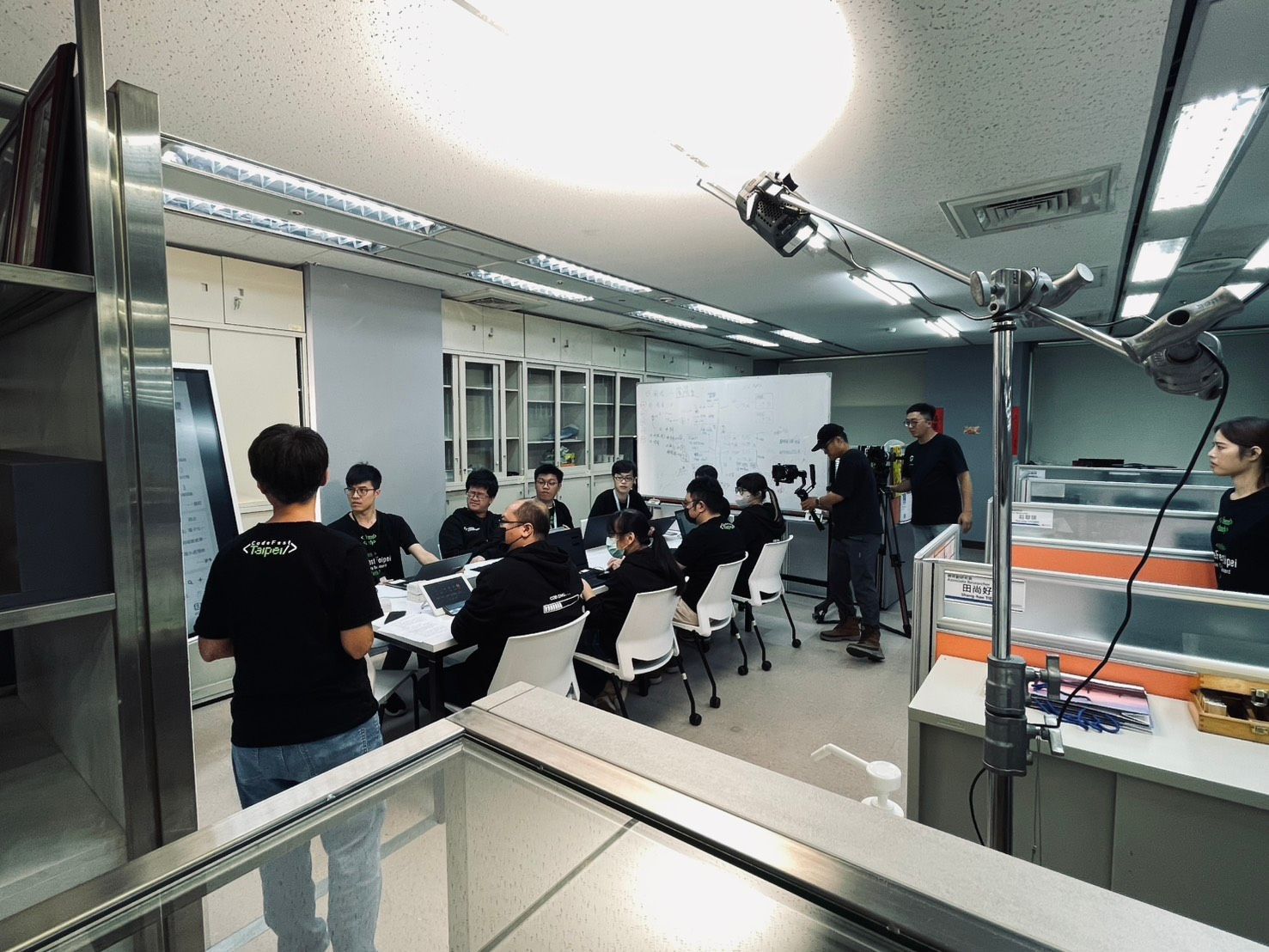一群人聚集在一間光線充足的辦公室裡,面前有一塊白板,圍坐在一張桌子旁;前面的人正在向他們講話。