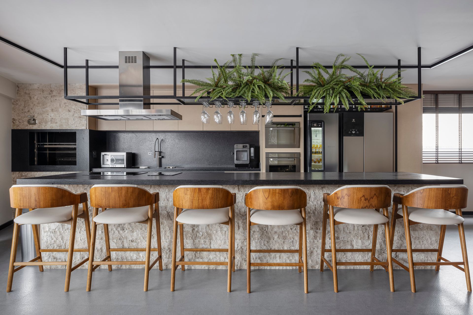 Cozinha moderna com ilha, seis banquetas e prateleiras suspensas com plantas. Bancadas escuras e eletrodomésticos pretos.