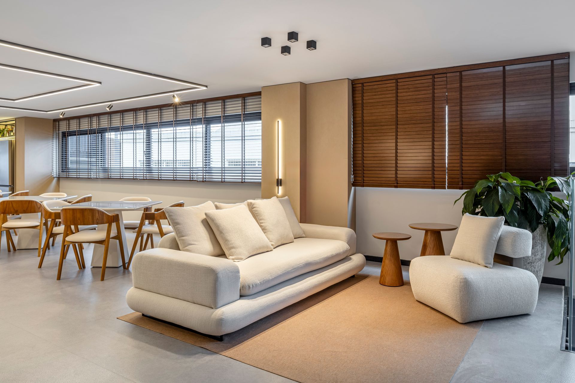 Sala de estar moderna com sofá branco, poltrona e mesas laterais de madeira. Inclui uma área de jantar e uma grande janela.