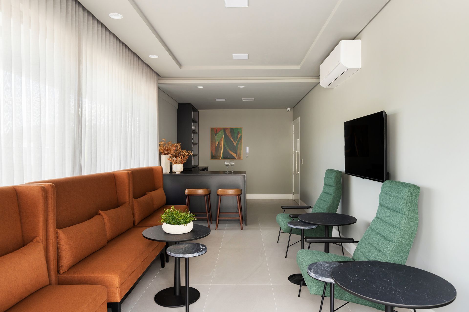 Sala de espera moderna com sofá laranja, cadeiras verdes e mesinhas. Um bar embutido está ao fundo.