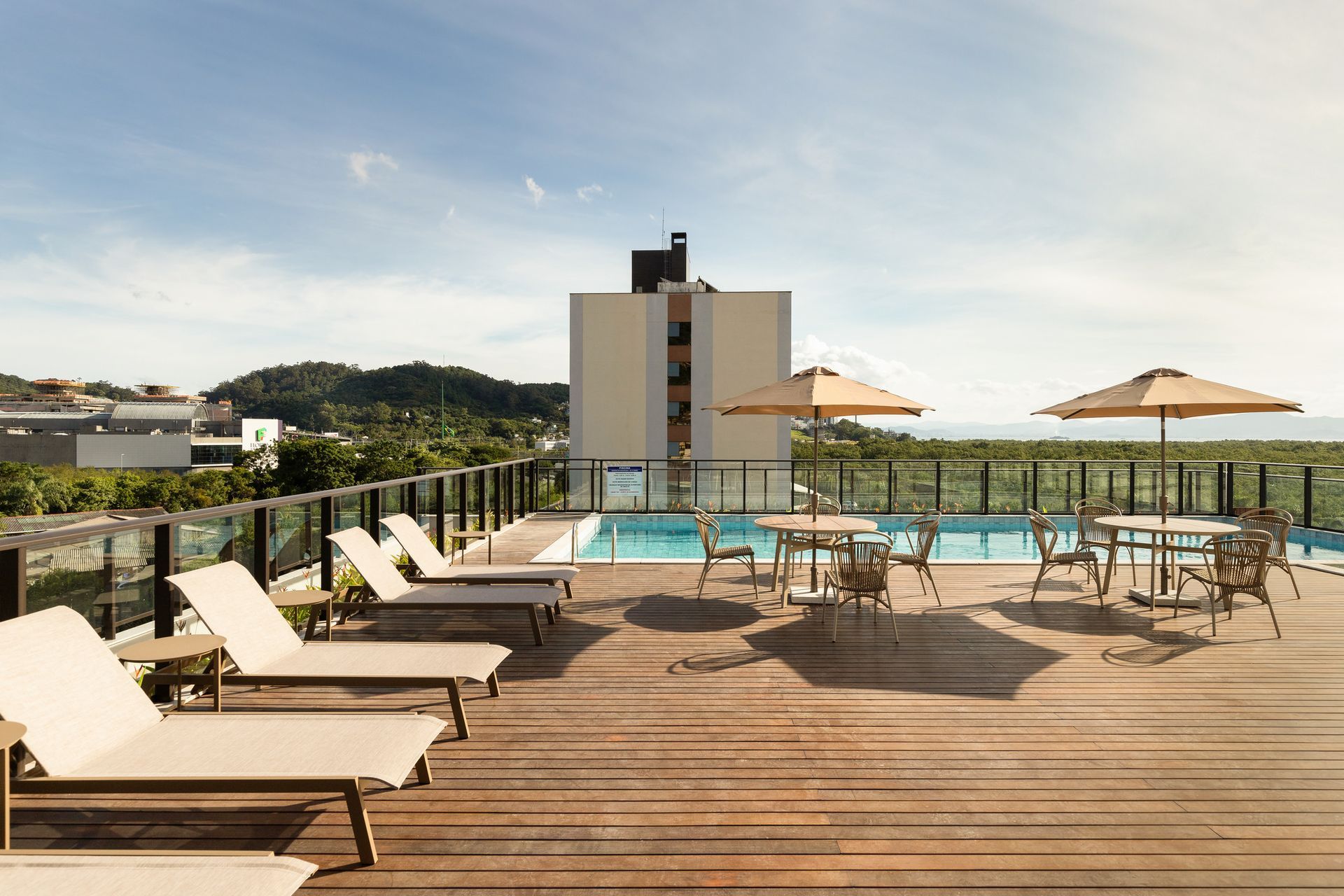Terraço com piscina na cobertura, espreguiçadeiras, mesas, guarda-sóis e um prédio sob um céu claro.