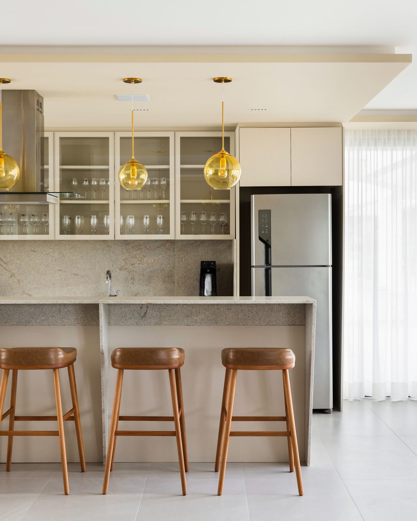 Cozinha com bancada de pedra, banquetas, armários de vidro e geladeira de aço inoxidável. Três luminárias pendentes douradas estão instaladas acima.