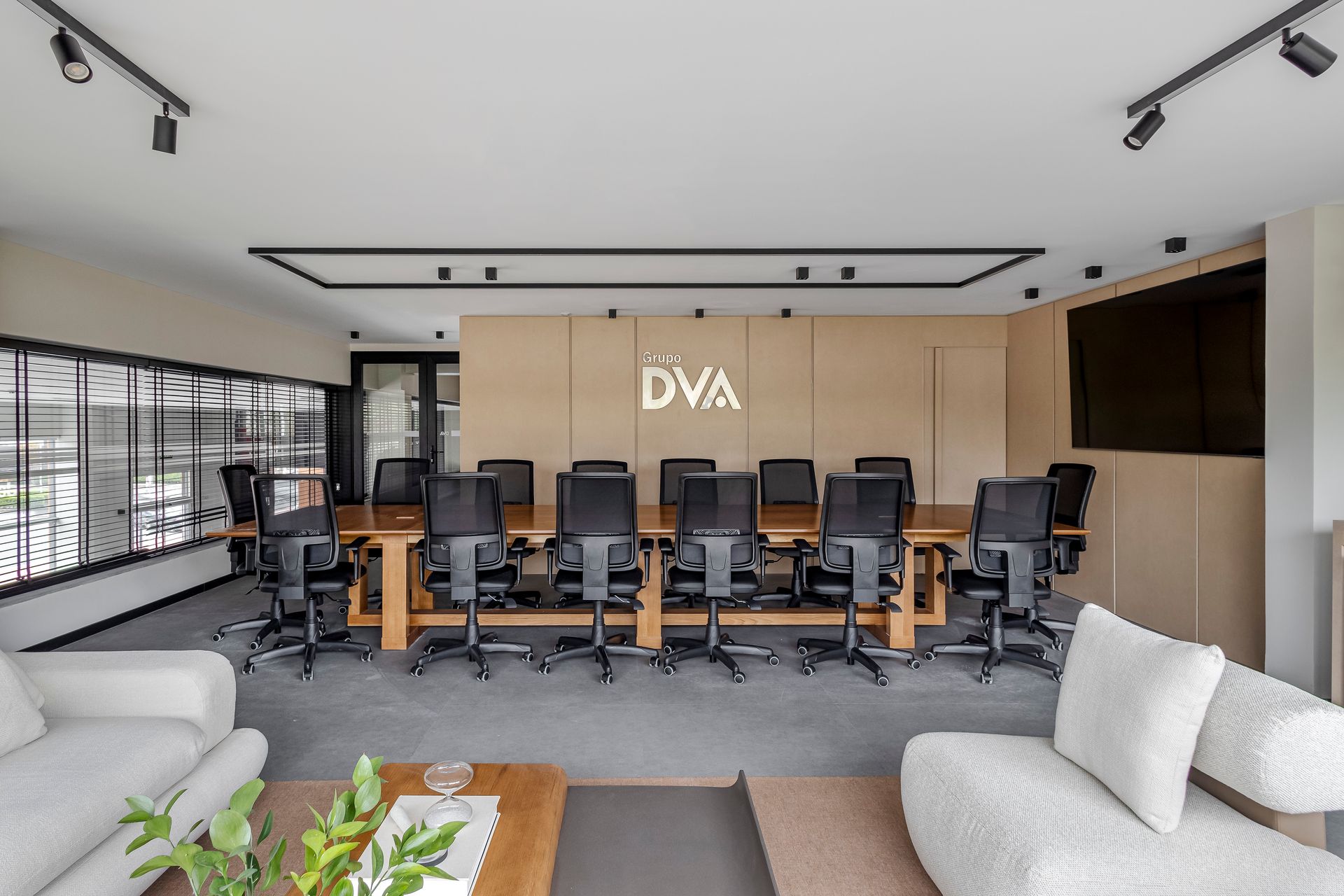 Sala de conferências com mesa comprida de madeira, cadeiras pretas, logotipo na parede e sofás.