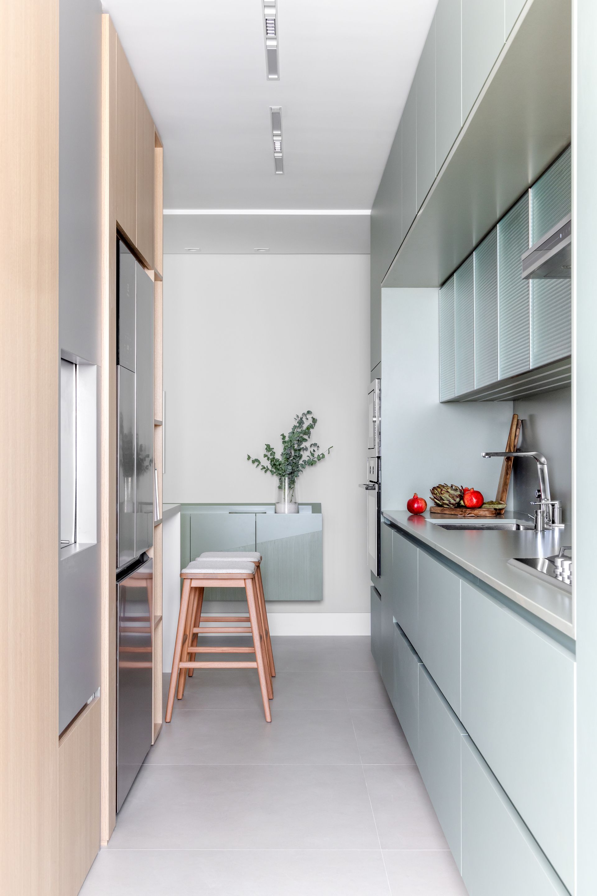 Cozinha estreita e moderna com armários azul-claros, eletrodomésticos de aço inoxidável e banqueta de madeira.